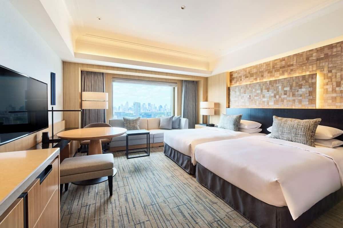 A hotel room with a city view, a flat-screen TV, a sofa, a dining table, and two beds.