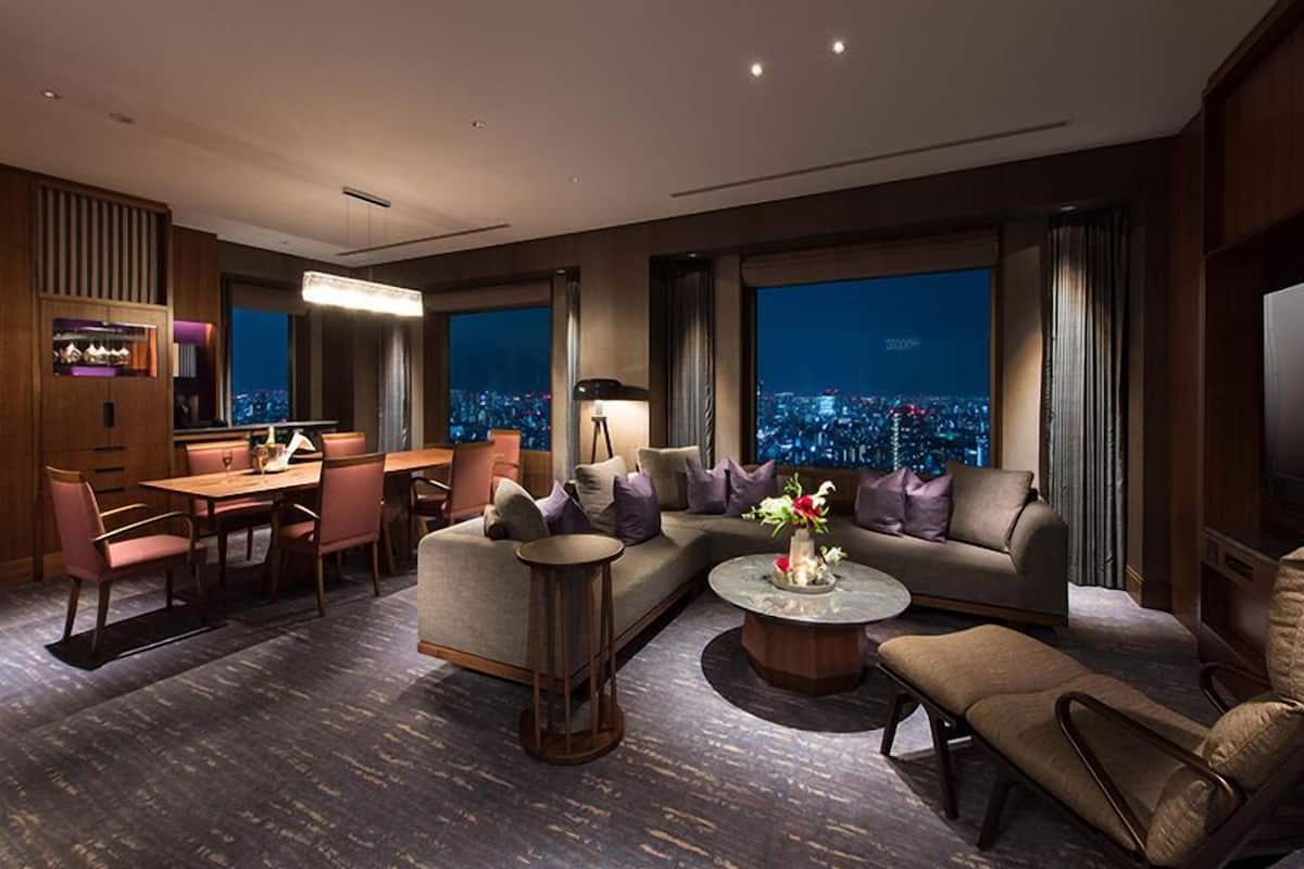 A modern hotel room with a dining area, a sofa set, a coffee table, and a city view through large windows.