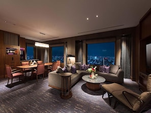 A modern hotel room with a dining area, a sofa set, a coffee table, and a city view through large windows.