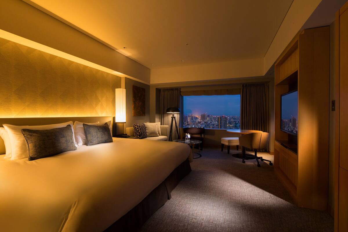 A hotel room with a large bed, a desk, a chair, and a view of the city at night.