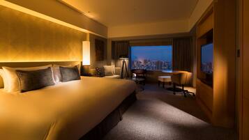 Habitación de hotel con una cama grande, un escritorio, una silla y vista a la ciudad de noche.