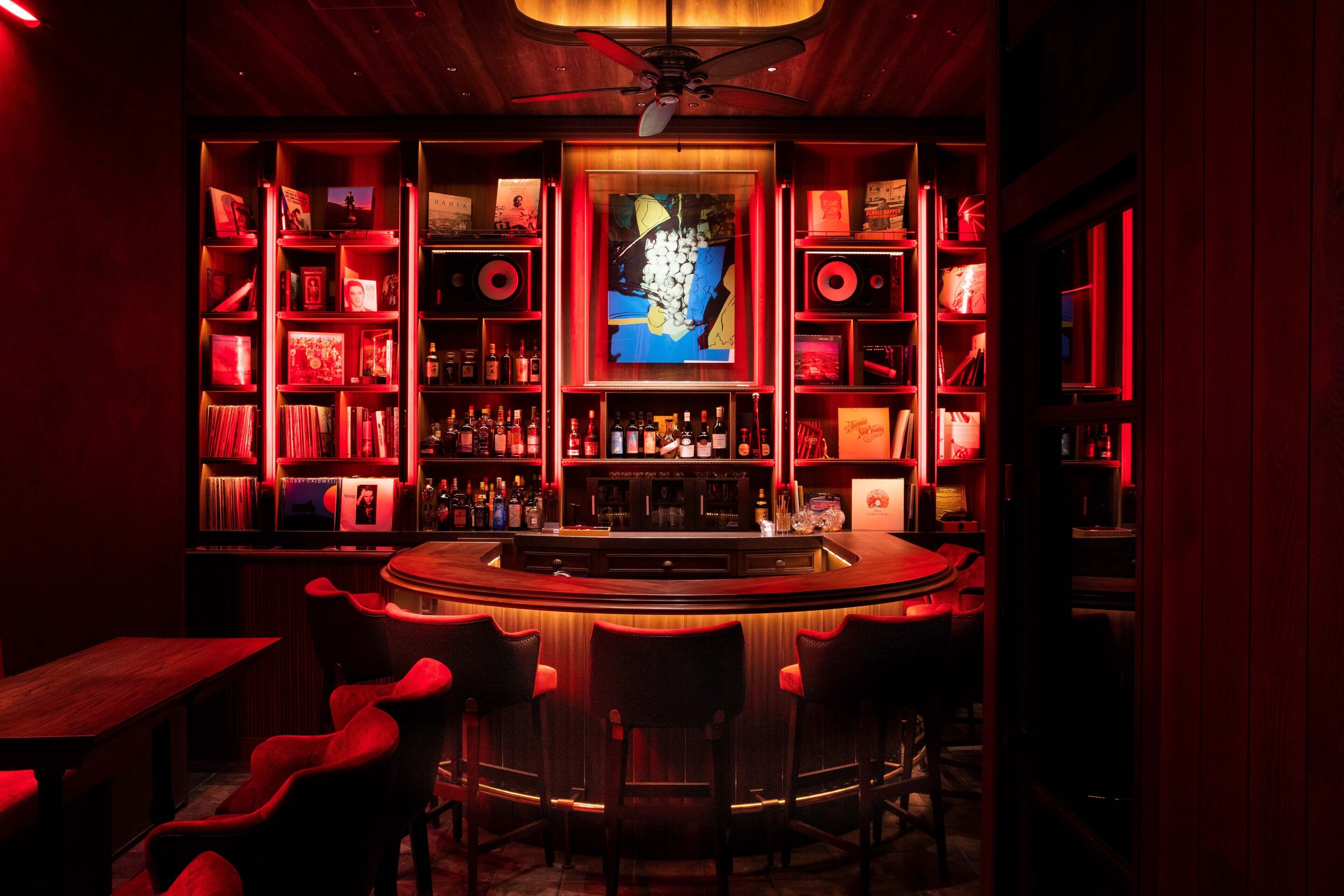 A bar area with a central counter, multiple shelves with bottles, and red upholstered chairs.