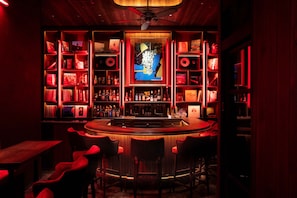 A bar area with a central counter, multiple shelves with bottles, and red upholstered chairs.