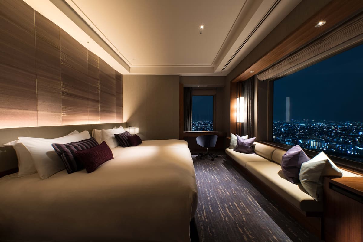 A hotel room with a large bed, a sofa, a desk, and a view of a cityscape at night.
