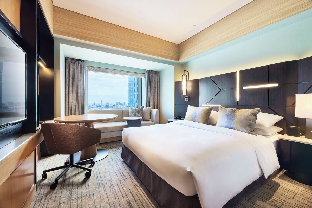 A modern hotel room with a large bed, a desk, a chair, and a view of the city.