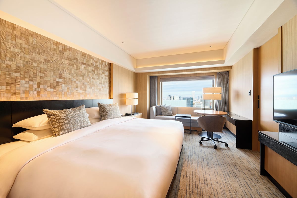 A hotel room with a large bed, a desk, a chair, a TV, and a city view.