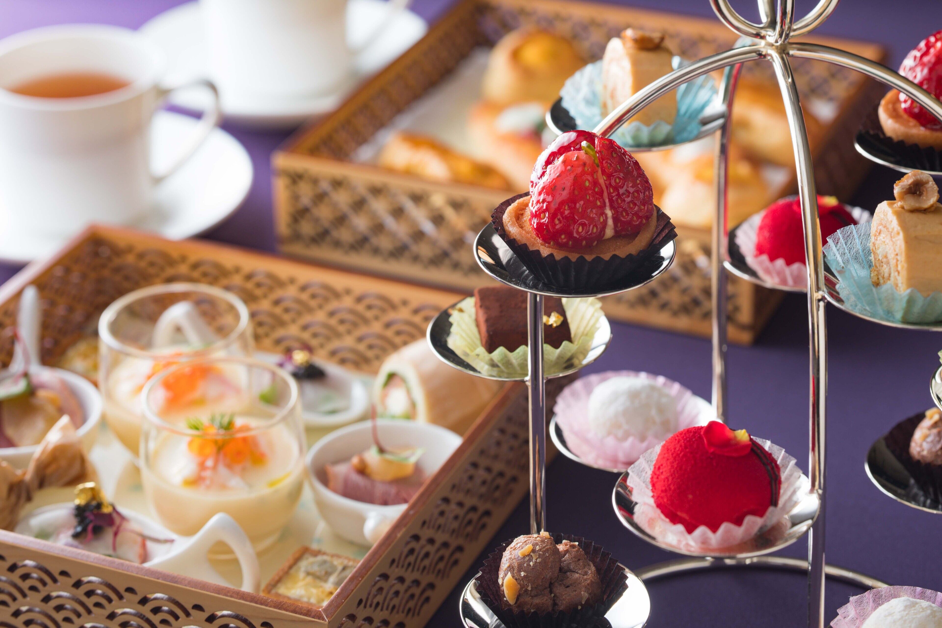 A variety of desserts on a multi-tiered stand, including cupcakes and a red dessert, with a cup of tea in the background.