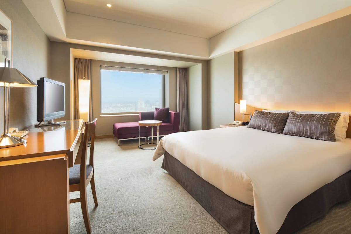 A hotel room with a large bed, a desk with a lamp, a chair, a small round table, and a television.