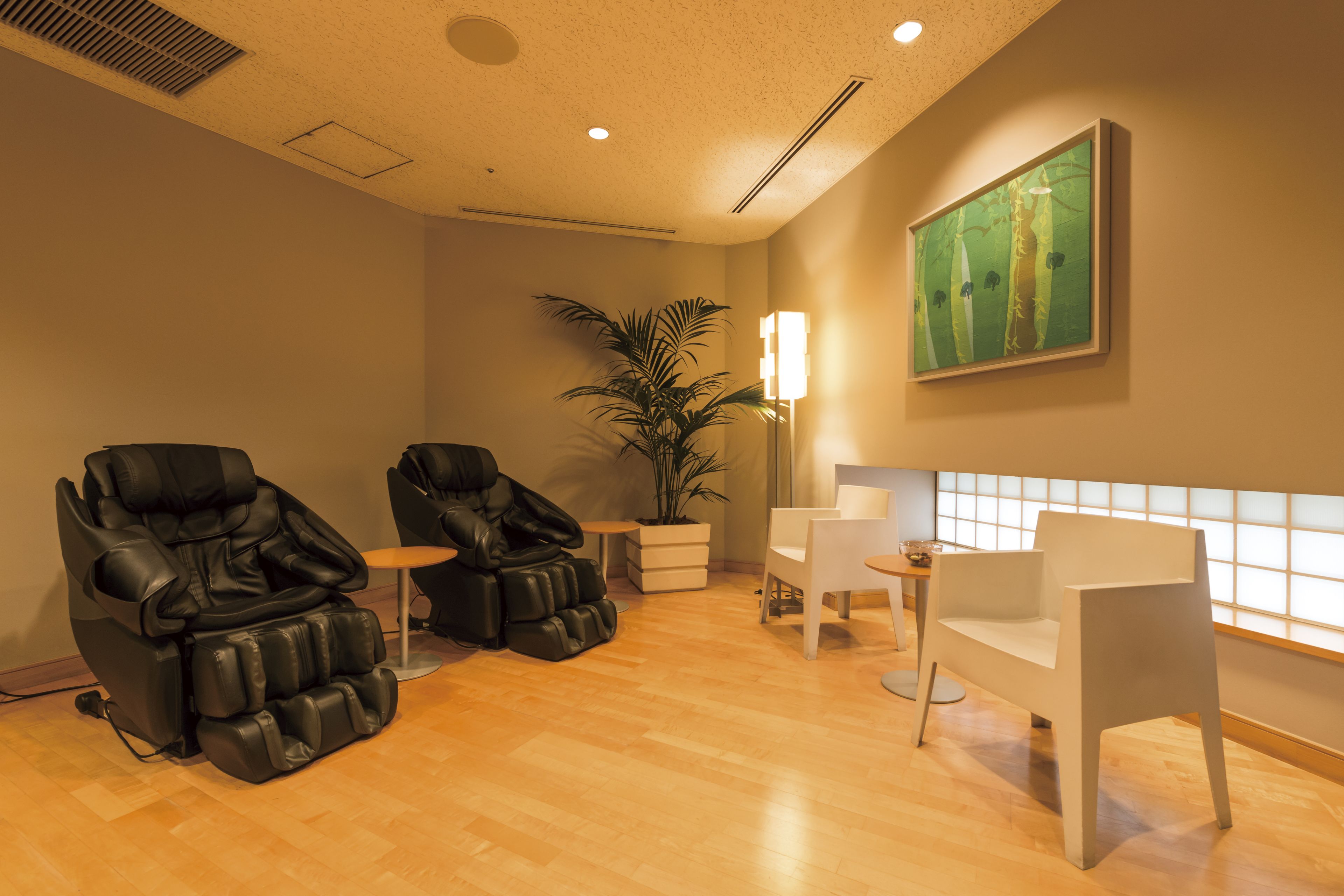 A massage therapy room with two massage chairs, a small table, a potted plant, and a wall-mounted painting.