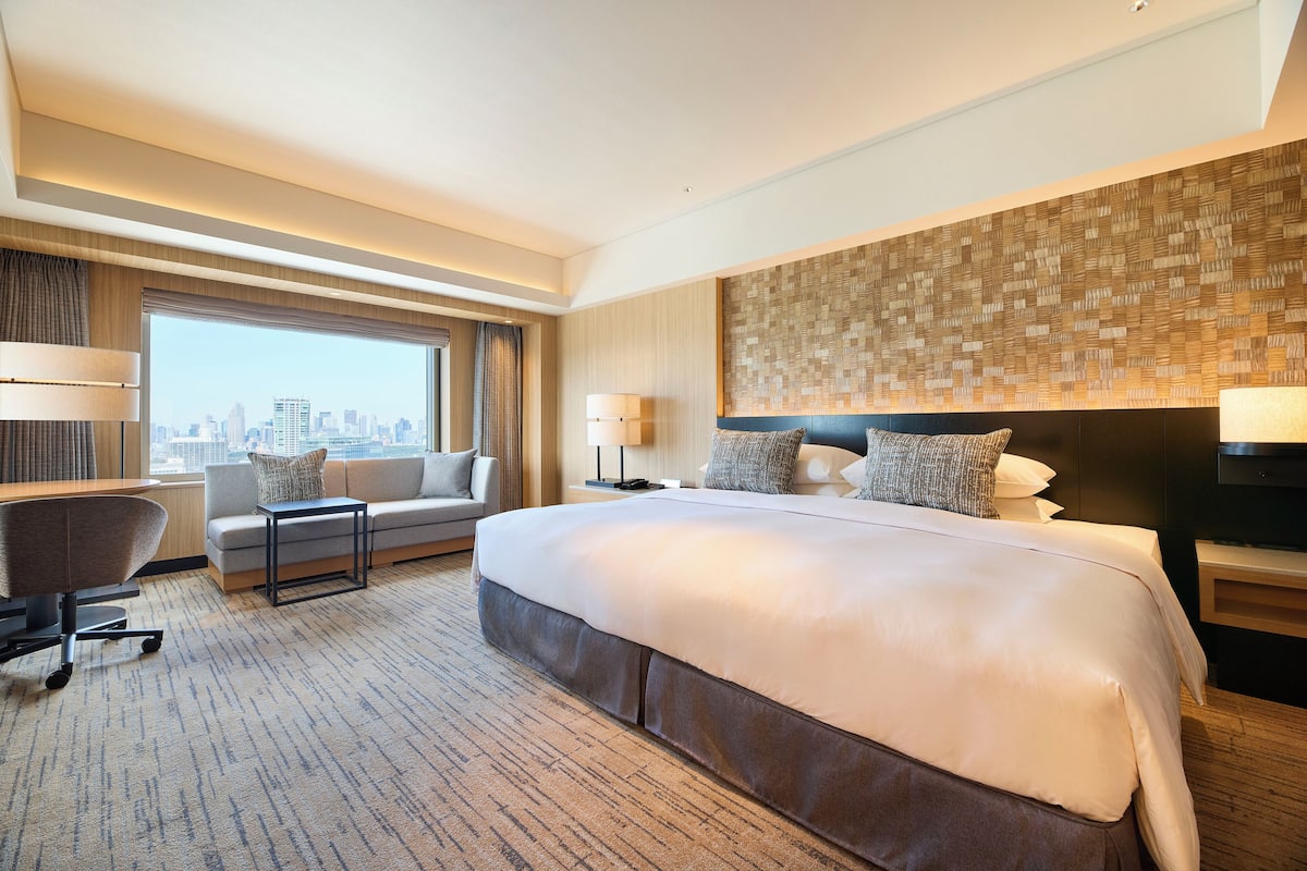 A modern hotel room with a large bed, a desk, a chair, and a view of the city skyline.