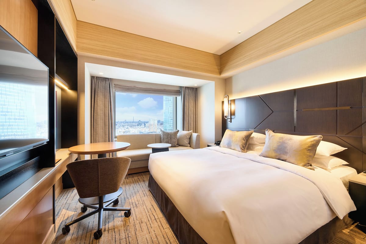 A hotel room with a large bed, a desk, a chair, a small table, and a view of the city.