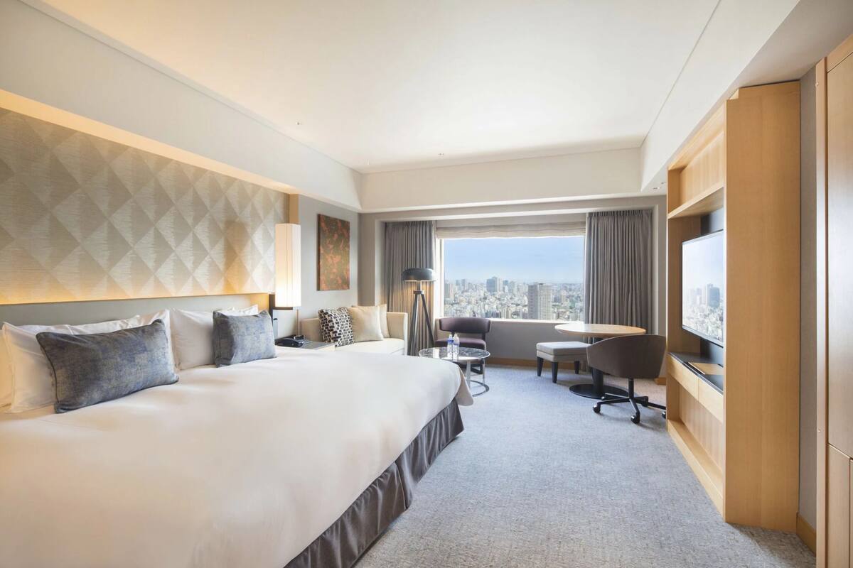 A hotel room with a large bed, a desk with a chair, a TV, and a view of the city.