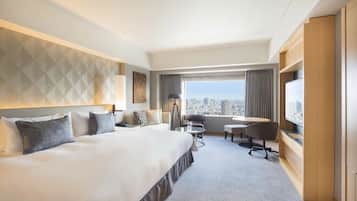 A hotel room with a large bed, a desk with a chair, a TV, and a view of the city.