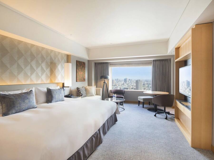 A hotel room with a large bed, a desk with a chair, a TV, and a view of the city.