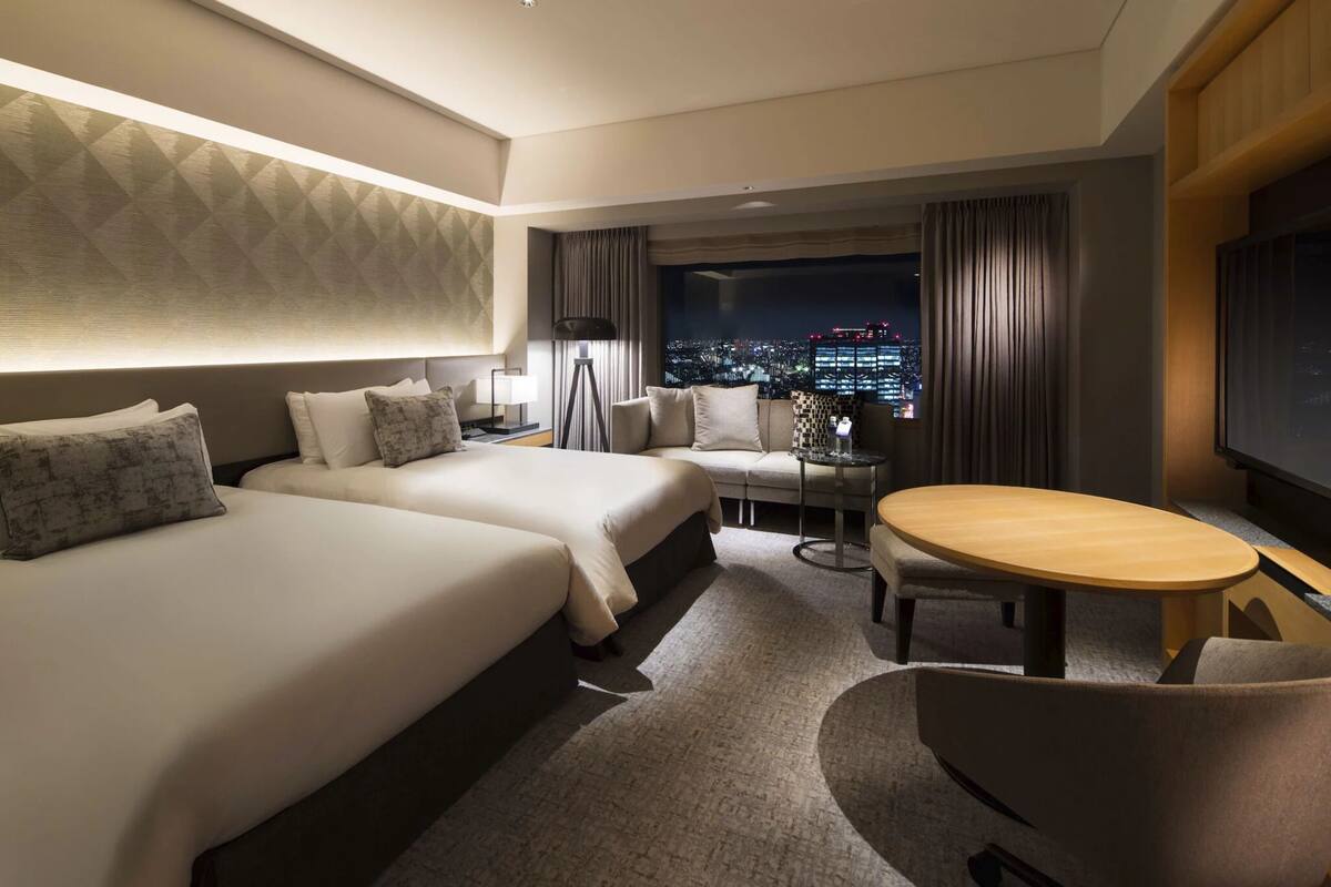 A hotel room with two beds, a small table, a chair, and a view of the city at night.