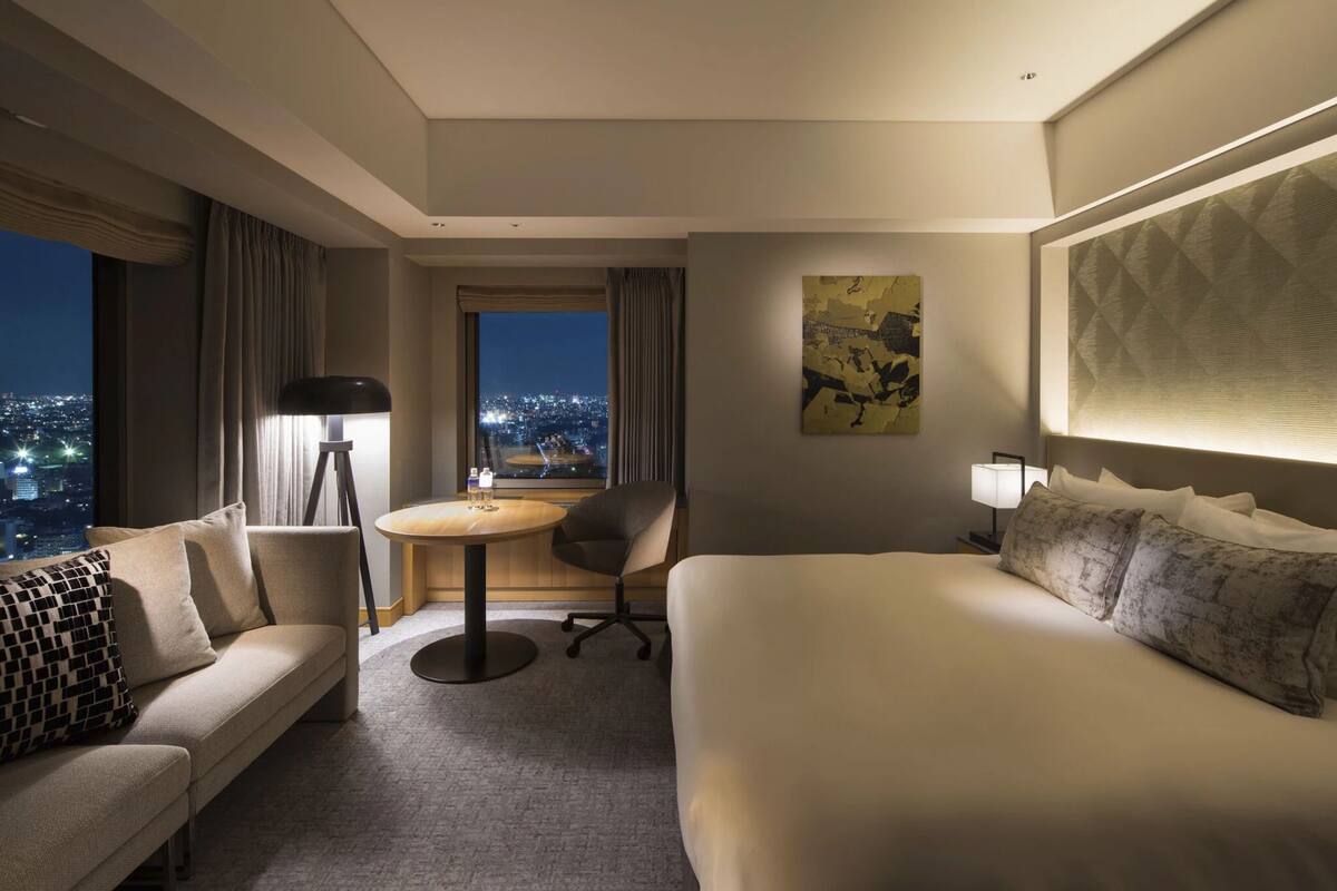 A hotel room with a bed, a sofa, a desk, and a chair. There is a window with a view of the city at night.