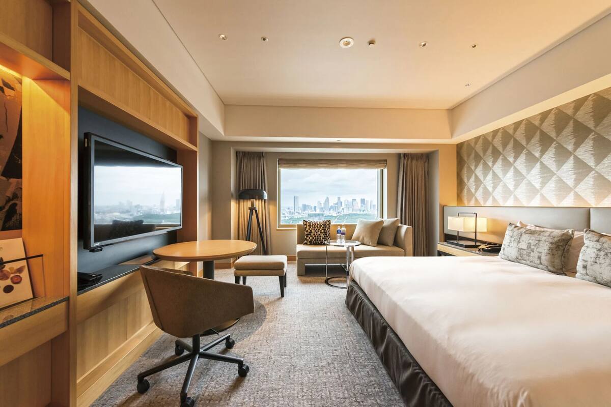 A hotel room with a large bed, a desk with a chair, a television, a sofa, and a view of a cityscape through a large window.
