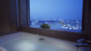 A modern bathroom with a large bathtub, a window with a city view, and a plant on the windowsill.