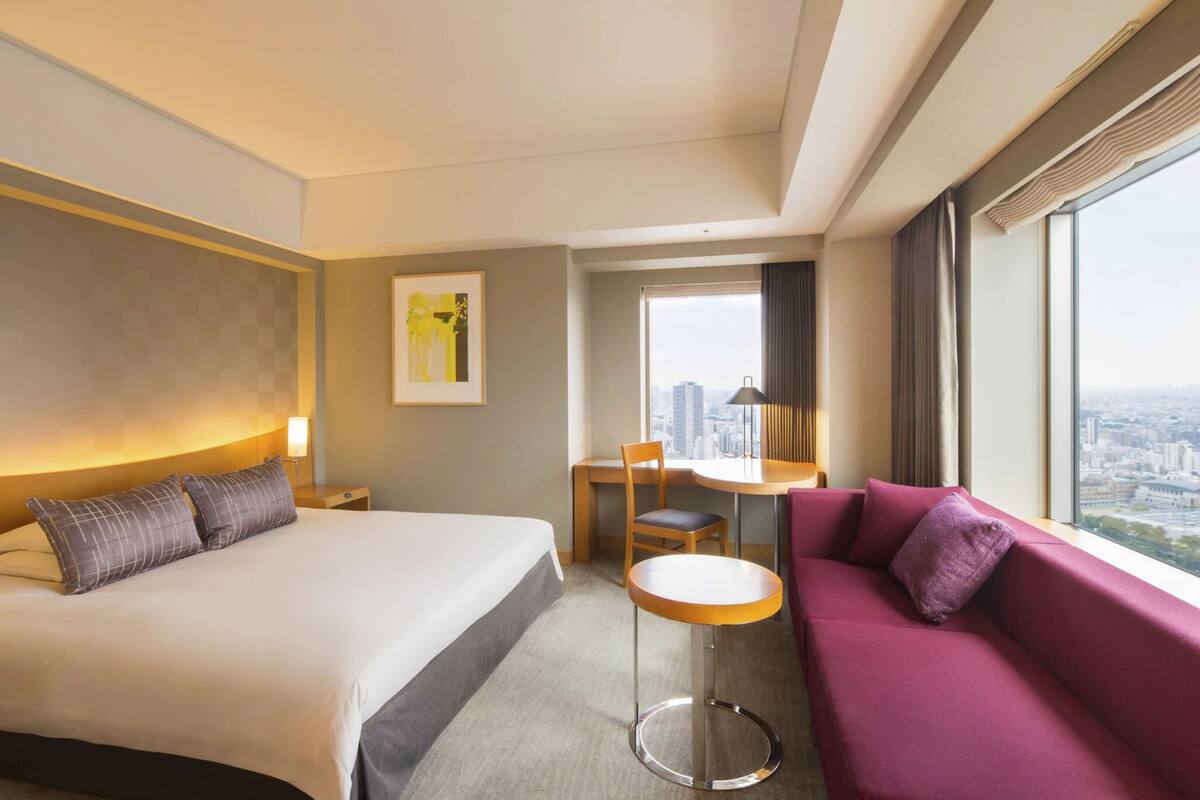 A hotel room with a bed, a small table, a chair, a sofa, and a large window with a city view.