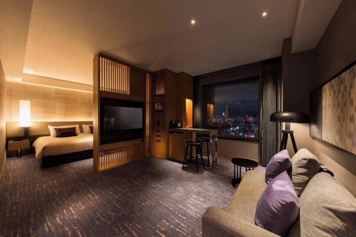 A modern hotel room with a large bed, a TV, a dining area, and a city view.