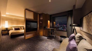 A modern hotel room with a bed, a TV, a dining area, and a large window with a city view.