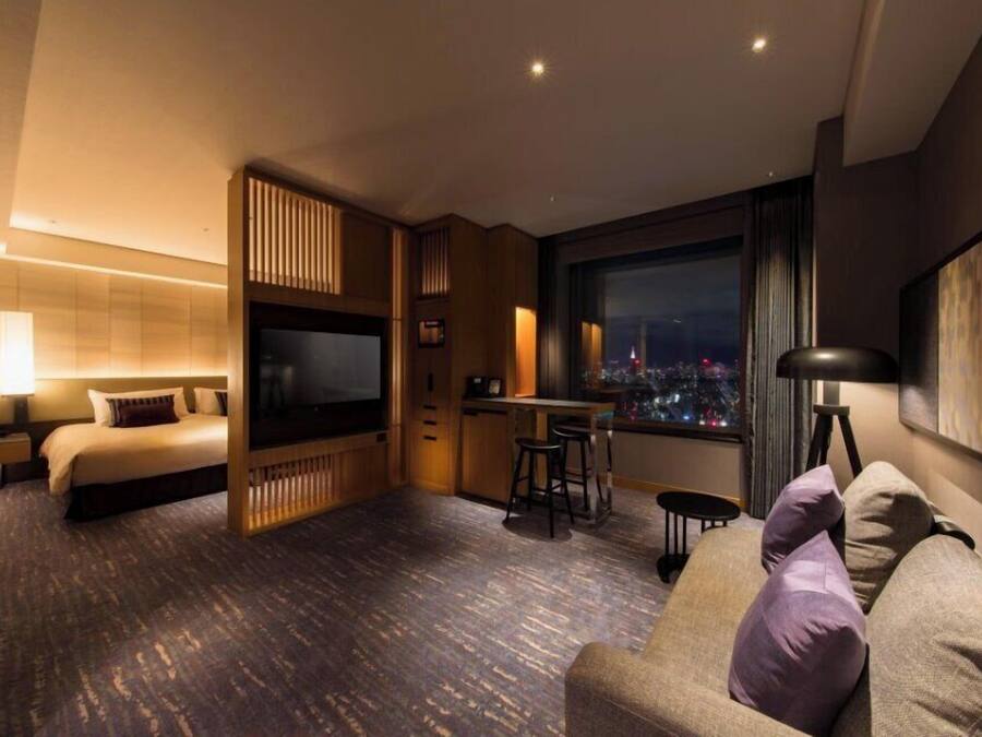 A modern hotel room with a bed, a TV, a dining area, and a large window with a city view.