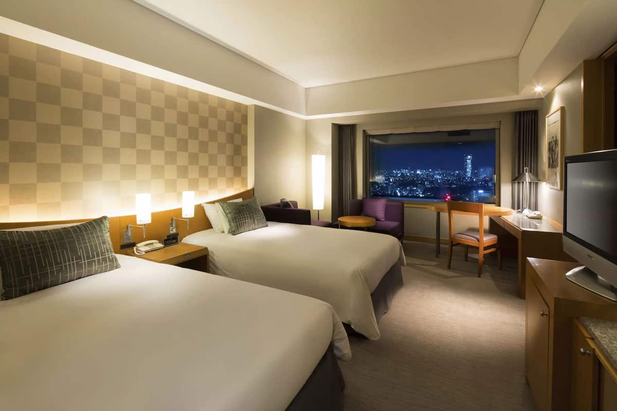 A hotel room with two beds, a desk, a chair, a television, and a window with a city view.