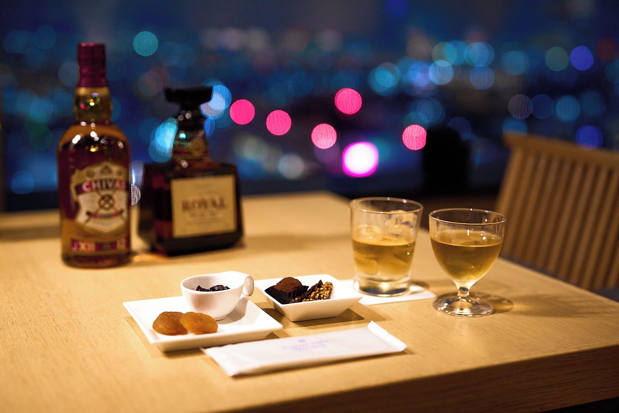 A table with a bottle of whiskey, a bottle of cognac, a glass of whiskey, a glass of water, and a plate with pastries.