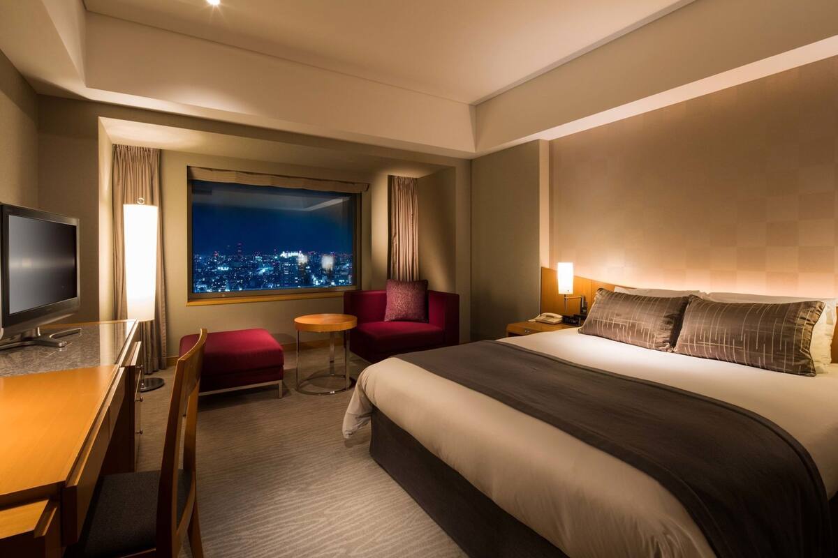 A hotel room with a bed, a desk, a TV, a chair, a small table, and a window with a city view.