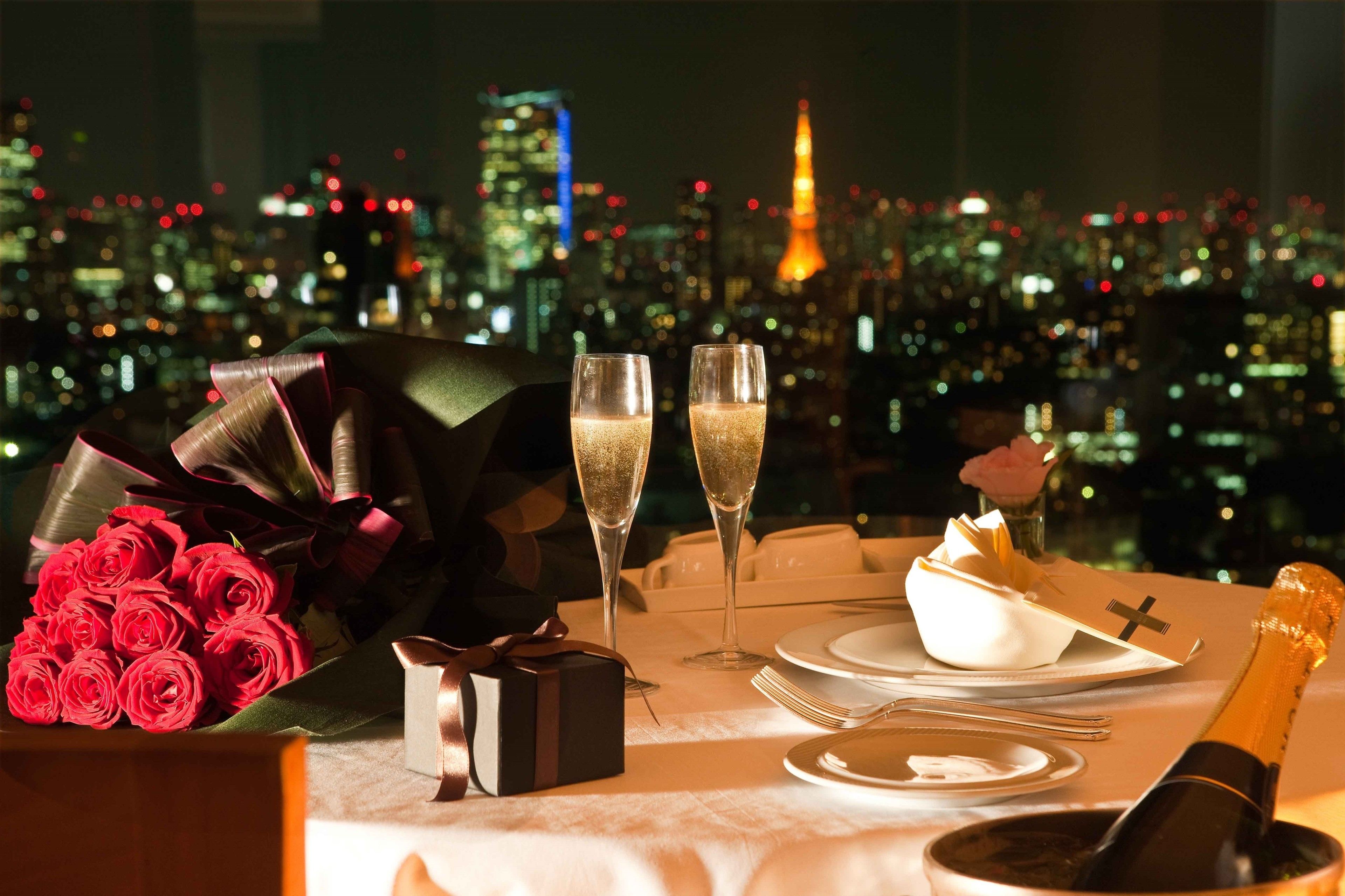 Ein romantisches Dinner mit Rosenblumen, Champagner und einer Geschenkschachtel auf einem Tisch, im Hintergrund ist die Stadt zu sehen.
