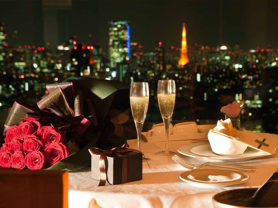 Ein romantisches Dinner mit Rosenblumen, Champagner und einer Geschenkschachtel auf einem Tisch, im Hintergrund ist die Stadt zu sehen.