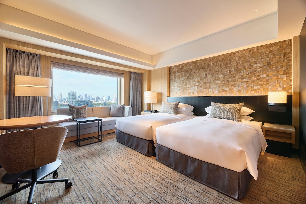 A hotel room with two beds, a desk, a chair, and a large window offering a city view.