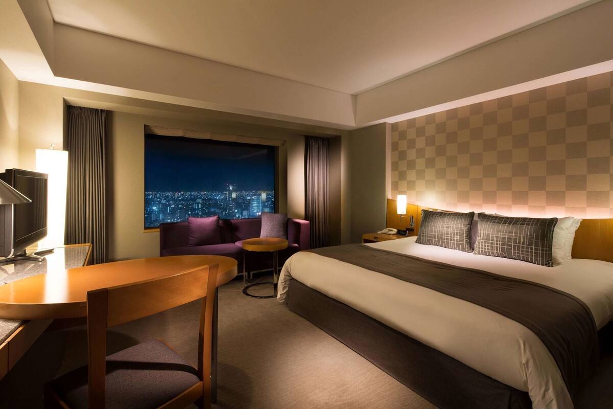 Habitación de hotel con cama, escritorio, silla, mesita, mesita de noche, lámpara, ventana con vista a la ciudad y un diseño de pared a cuadros.