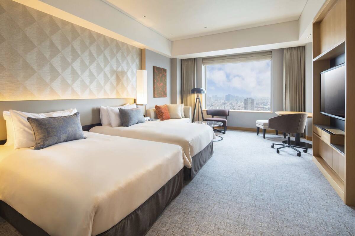 A hotel room with two beds, a desk, a chair, a TV, and a large window with a city view.