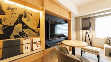 A hotel room with a flat-screen TV mounted on a wooden panel, a round table with two chairs, a bench, and a large abstract painting on the wall.