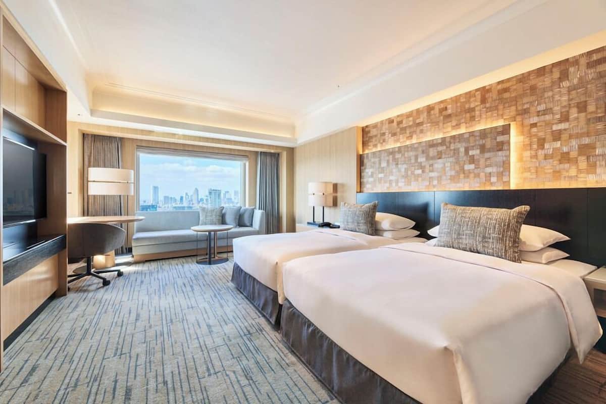 A hotel room with two beds, a TV, a sofa, a desk, and a view of the city.