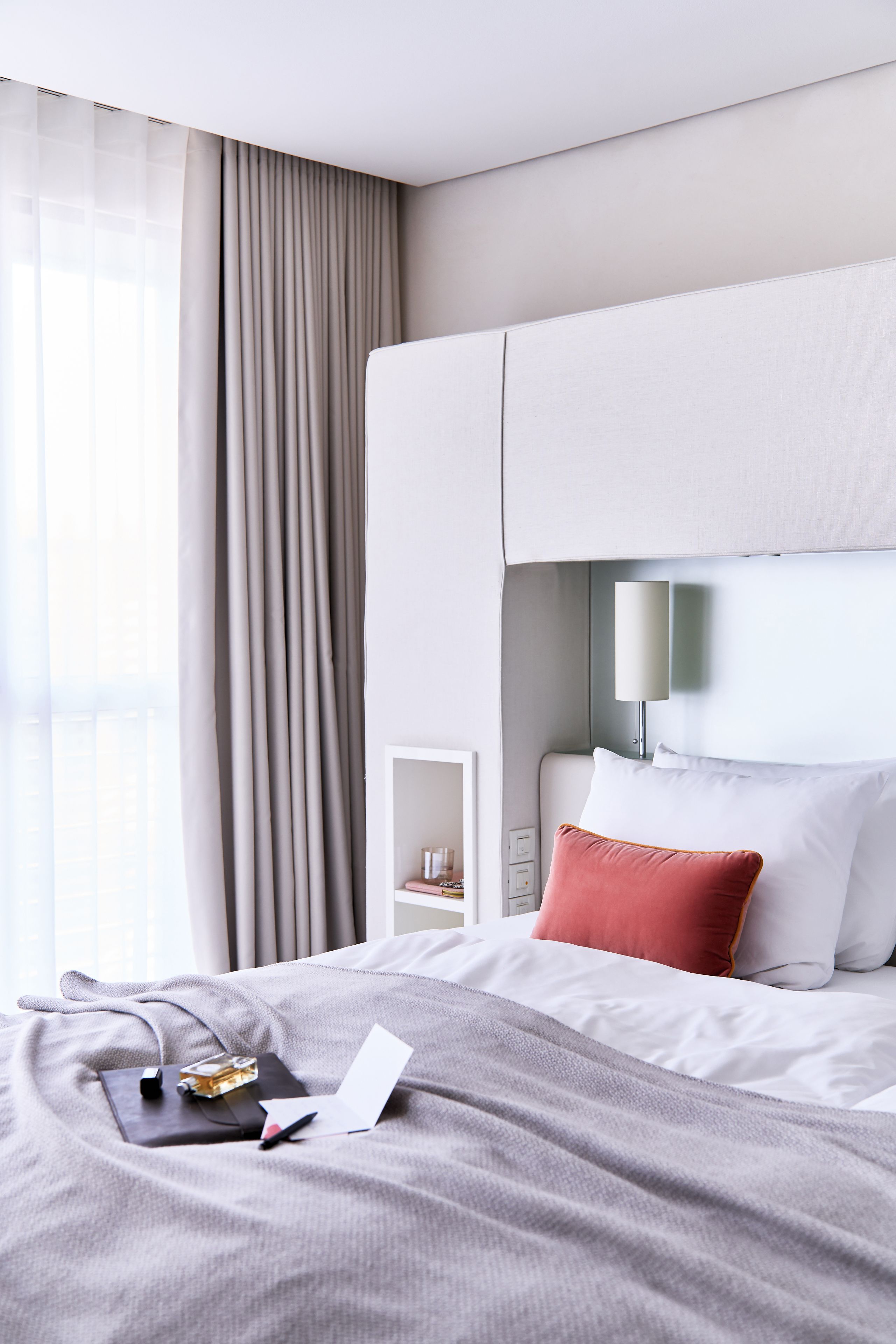 A modern hotel room with a neatly made bed, a bedside table with a lamp, and a view of the city through large windows.