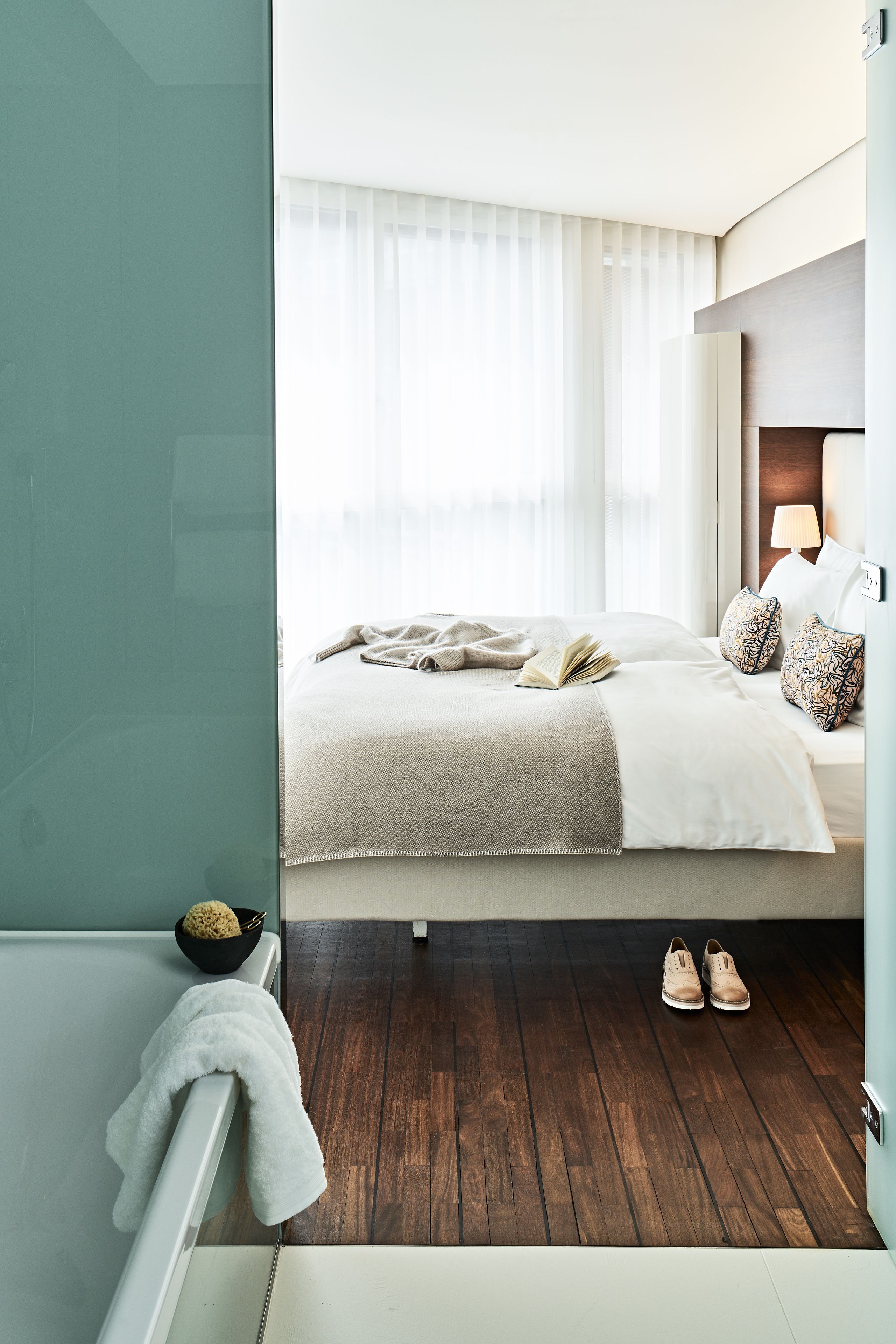 A modern hotel room with a large bed, a bedside table with a lamp, and a view of the city through sheer curtains.