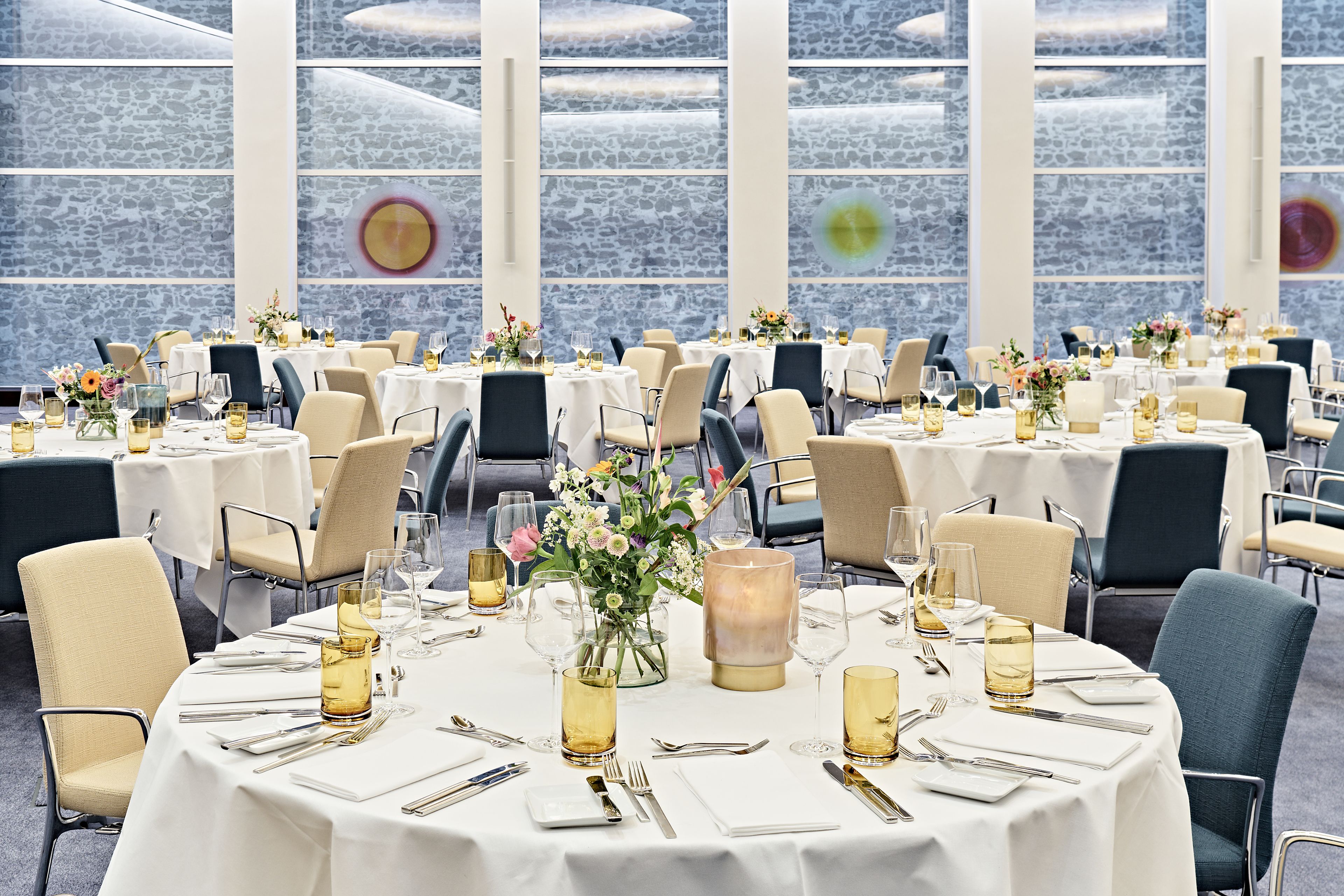 A spacious dining area with round tables set for a formal meal, featuring white tablecloths, elegant chairs, and floral centerpieces.