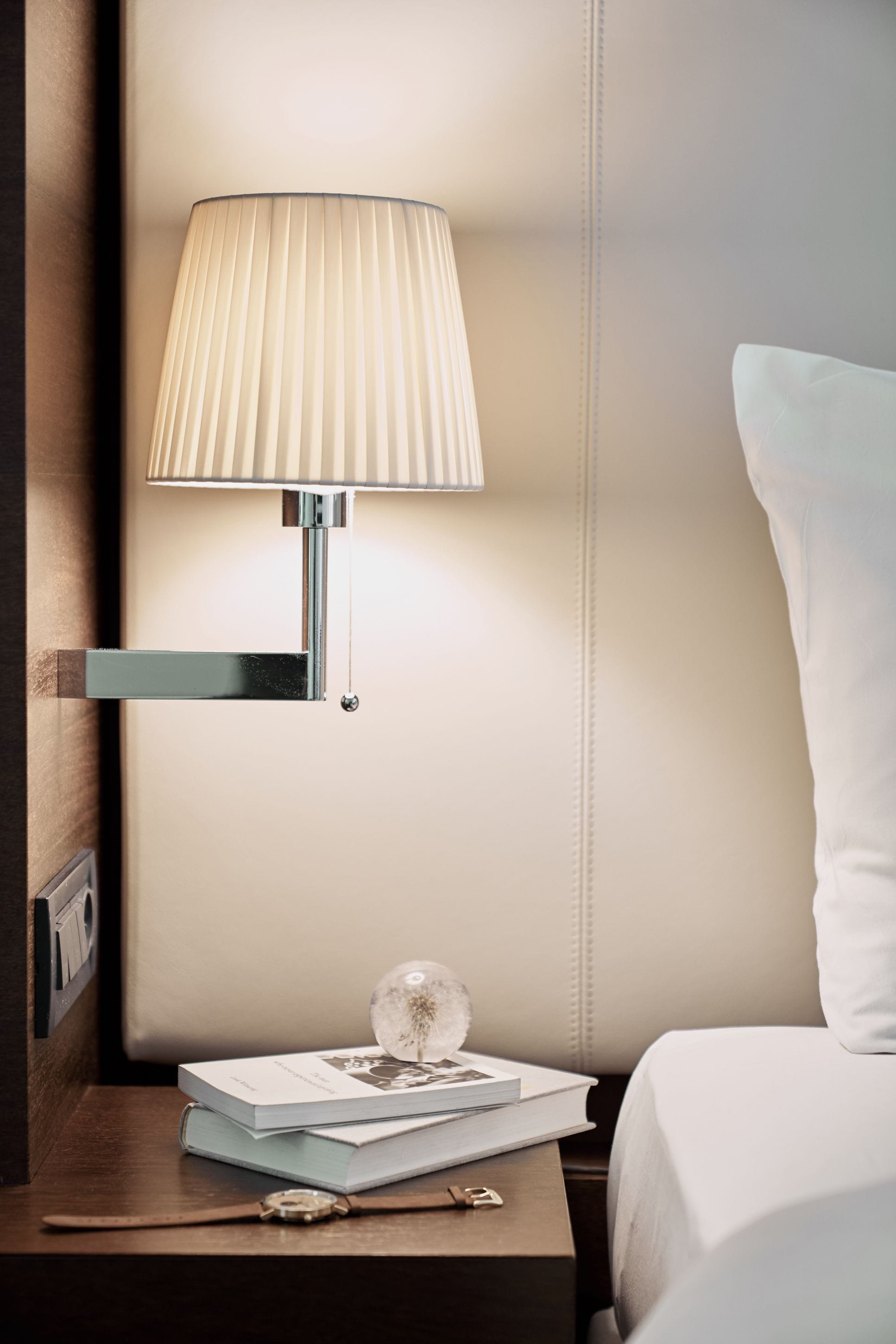 A neatly made bed with a white pillow, a bedside table with a lamp, a book, and a watch.