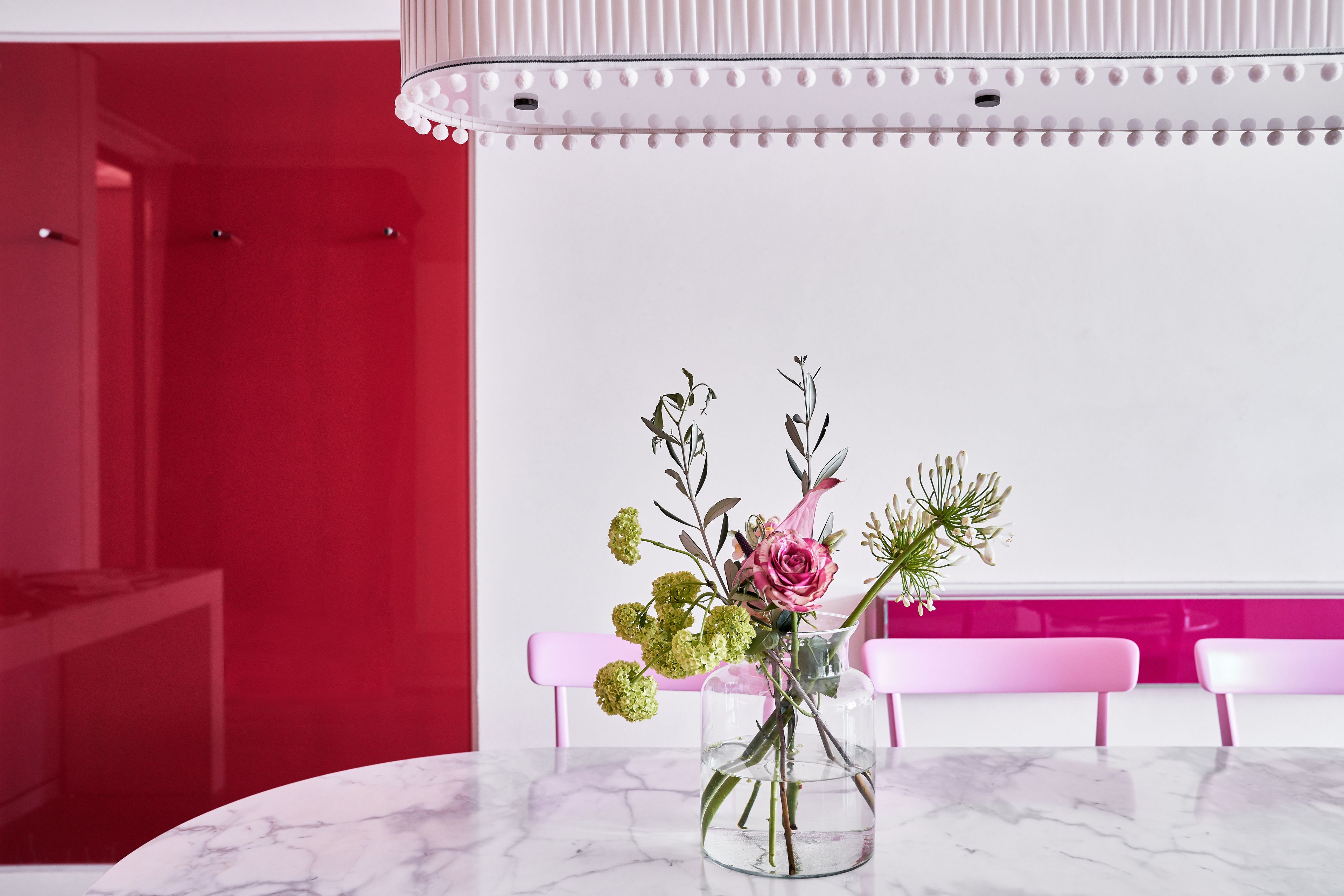 Ein Marmortisch mit einer klaren Vase, die Blumen enthält, rosafarbene Stühle und eine rote Wand.