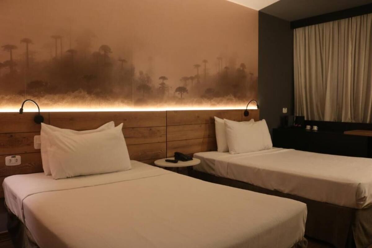 Habitació d'hotel amb dos llits individuals, una taula rodona petita i un panell il·luminat integrat al capçal del llit.