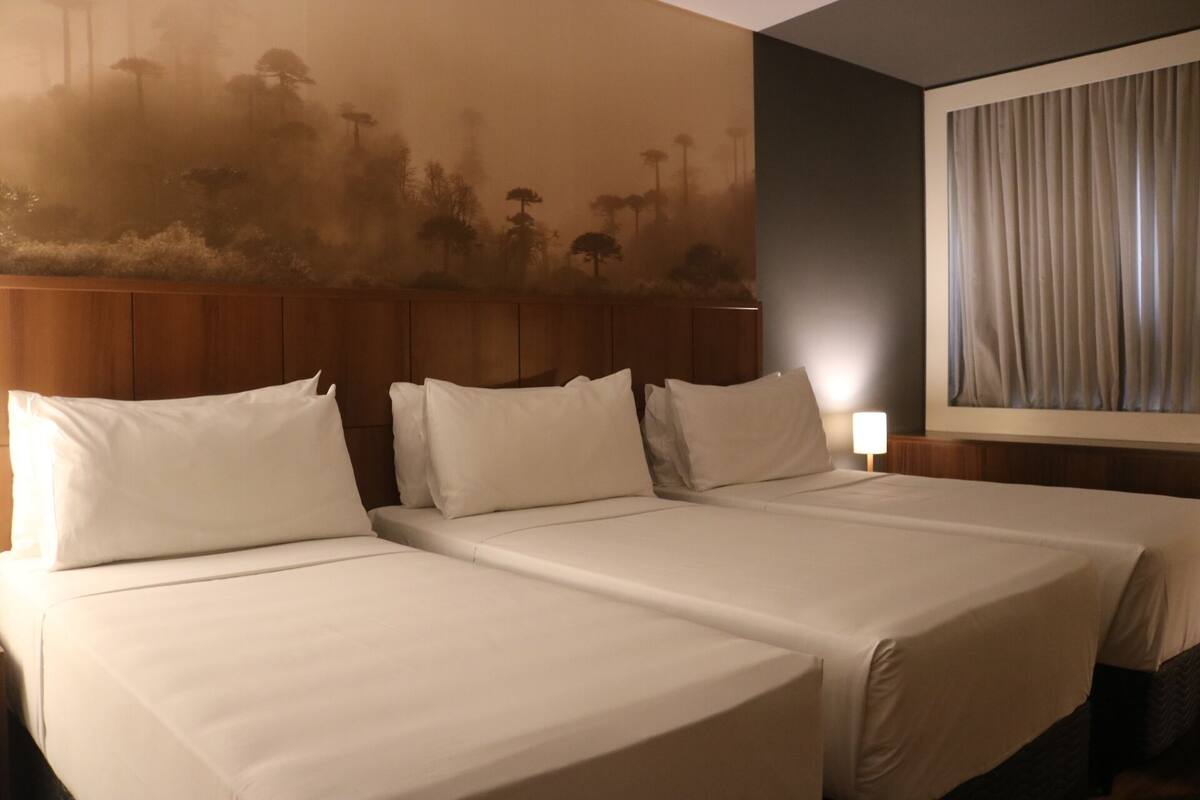 Habitació d'hotel amb dos llits, un panell de fusta al capçal, un quadre gran amb arbres i una finestra amb cortines.