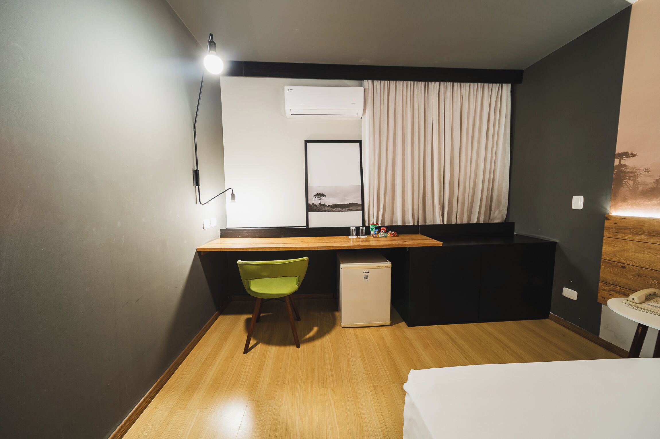 Une chambre d’hôtel compacte comprenant un bureau en bois, un petit réfrigérateur, une chaise verte, un lit et une table de chevet avec un téléphone.