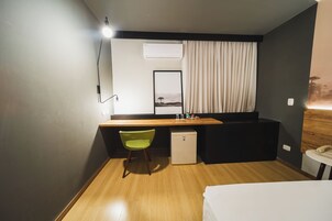 Habitación de hotel pequeña y funcional con escritorio de madera, refrigerador, silla verde, cama y mesita de noche con teléfono.