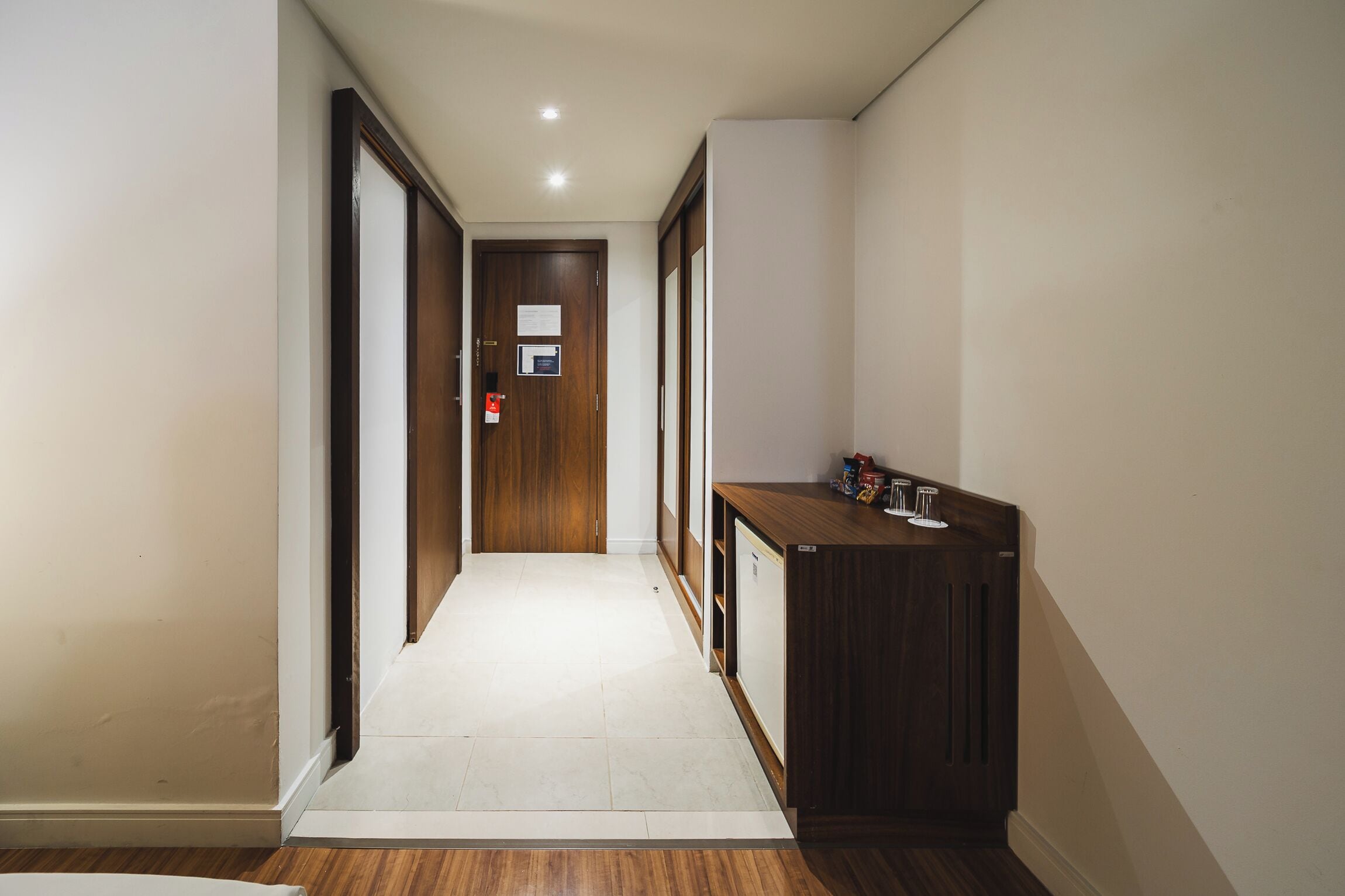 Un pasillo estrecho con puertas de madera, suelo de baldosas blancas y un pequeño mostrador de madera con algunos objetos encima.