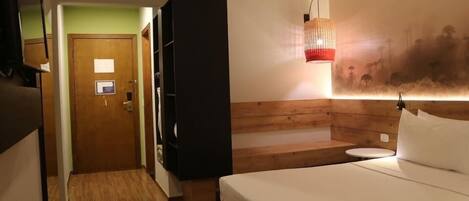 A hotel room with a bed, a wooden headboard, a nightstand, a wall-mounted lamp, and a door leading to another room.