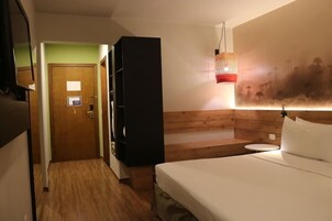Una habitación de hotel con una cama, cabecera de madera, mesita de noche, lámpara fijada a la pared y una puerta que da a otra habitación.