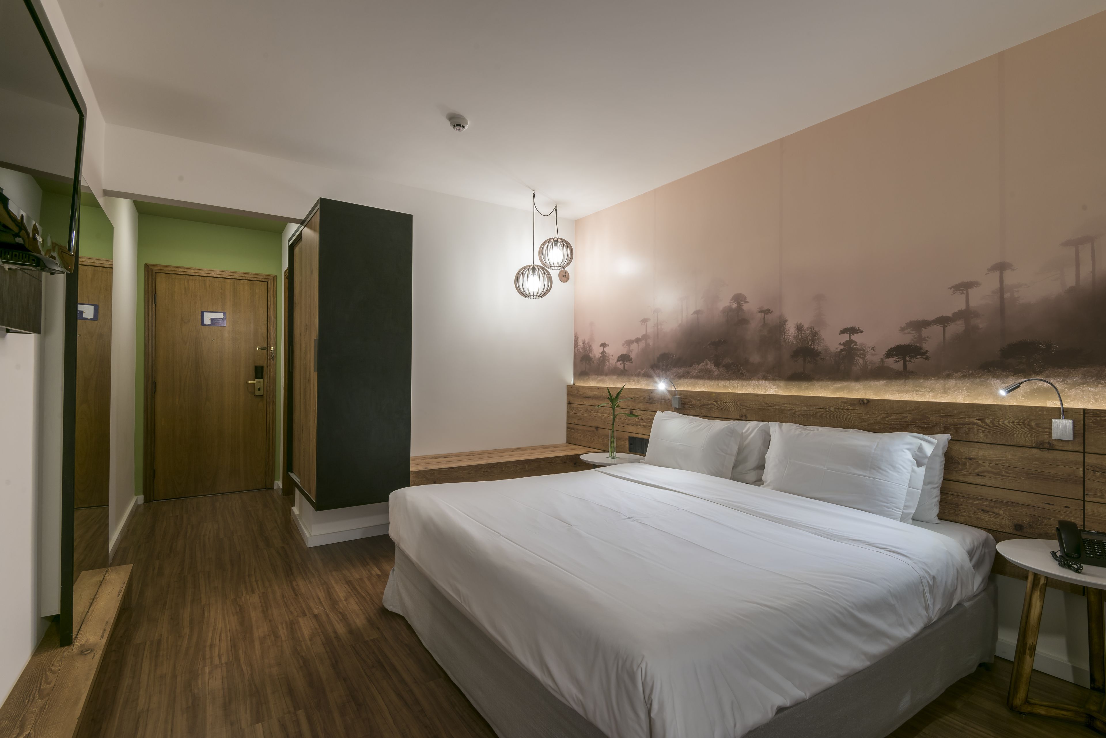 A hotel room with a large bed, a wooden headboard, a nightstand, a wall-mounted phone, and a painting of trees.
