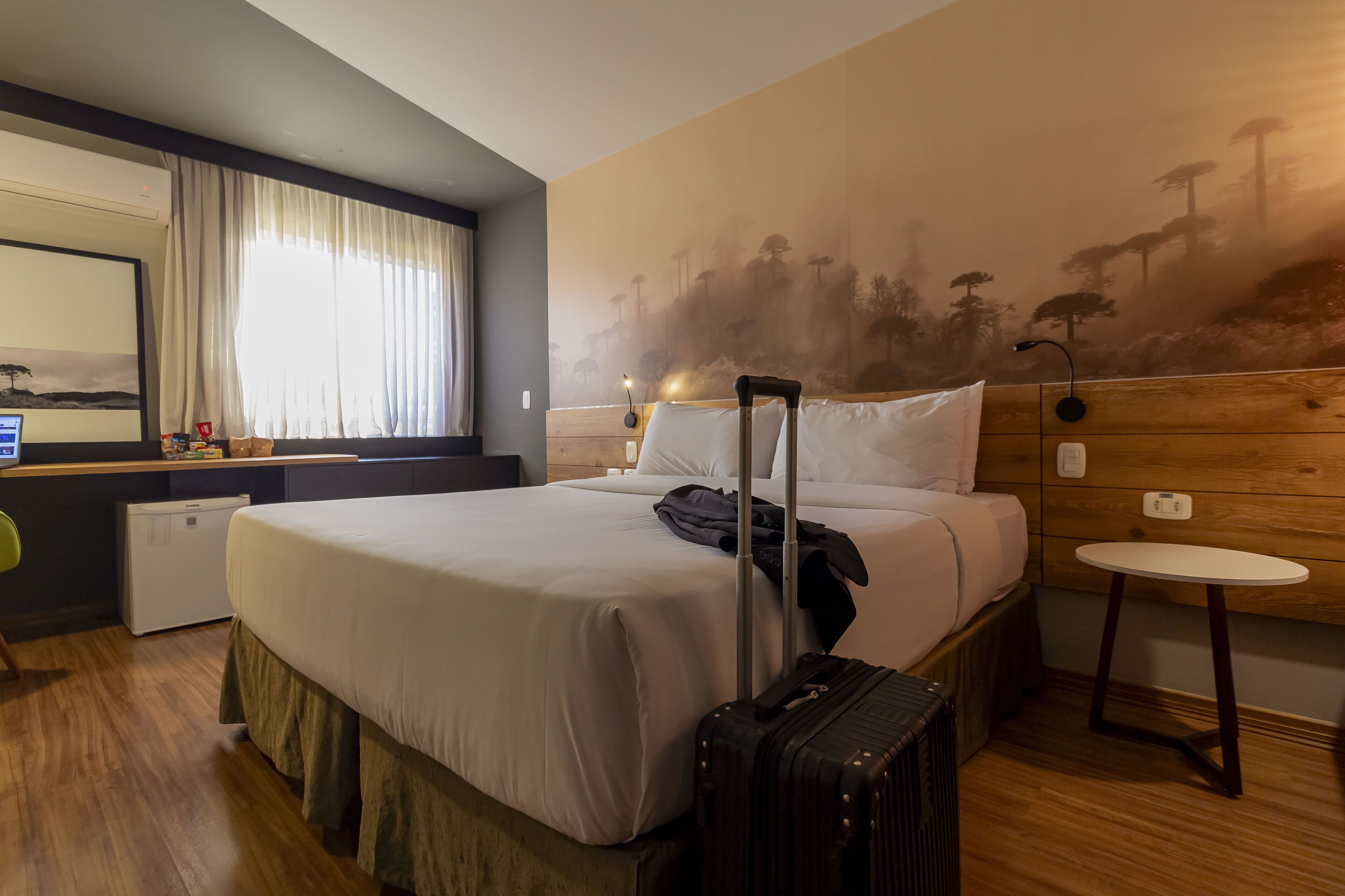 Quarto de hotel com cama, escrivaninha com computador, mini-geladeira e mala.